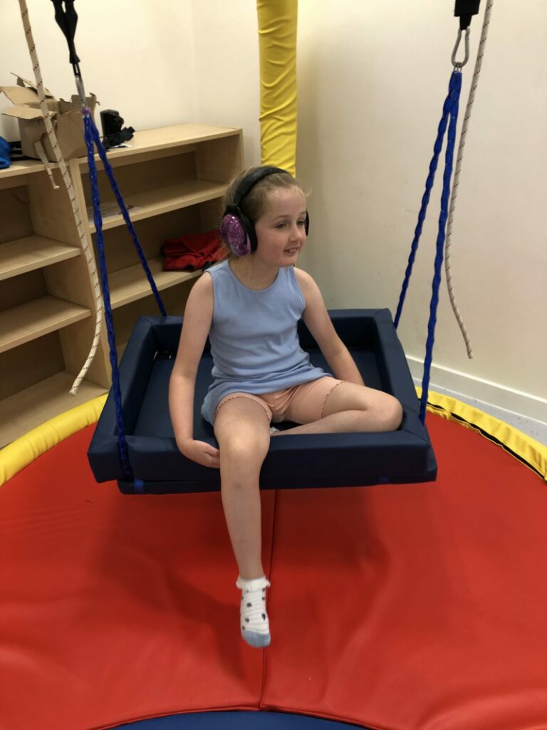 A little girl wearing headphones is sat on a platform swing