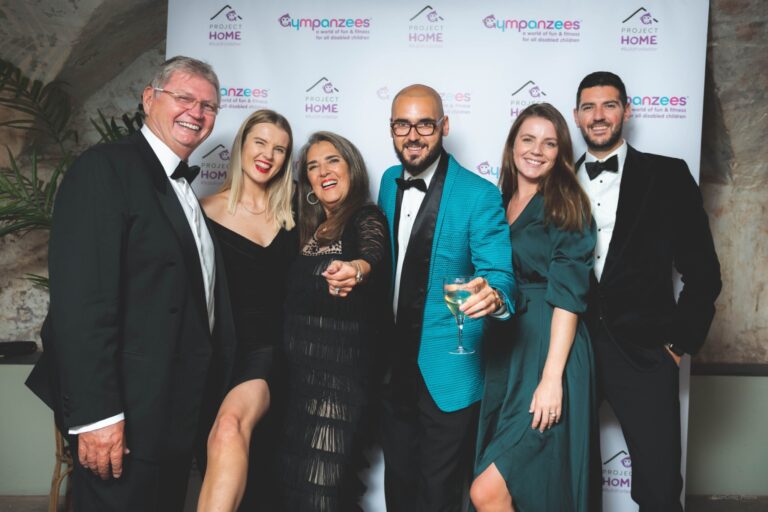 Six guests smile at the camera dressed in suits and glamorous dresses.