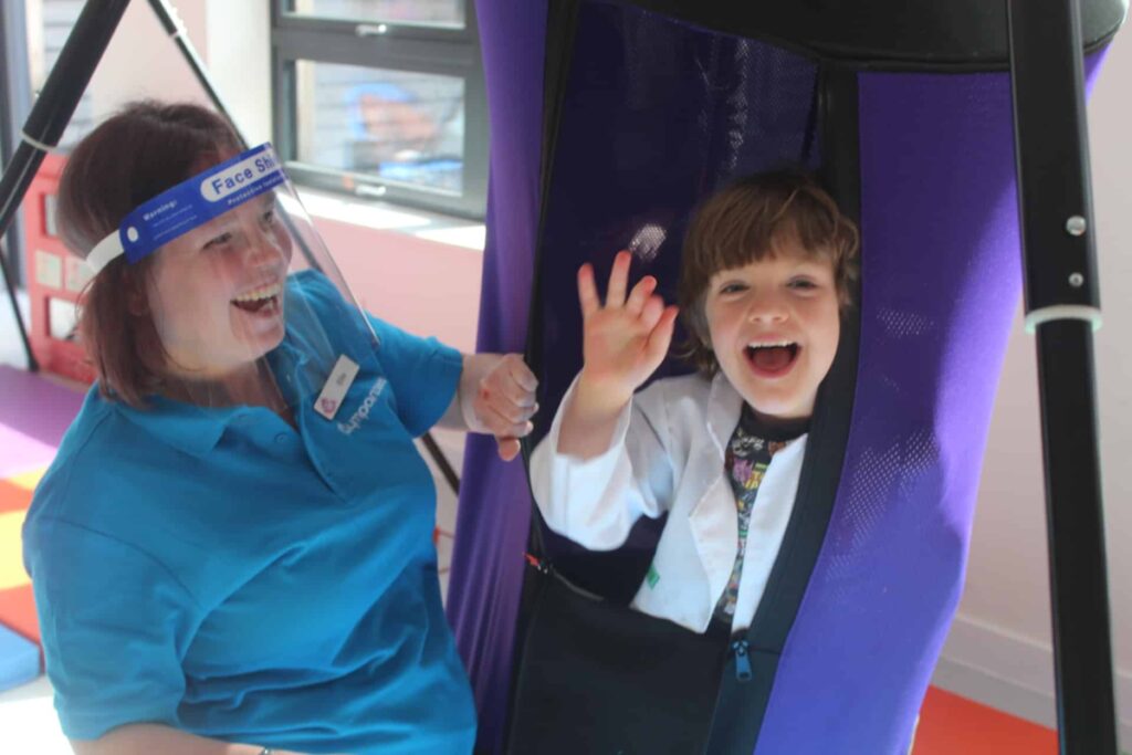 A small child is in a purple swing, next to them is an adult in a blue Gympanzees top