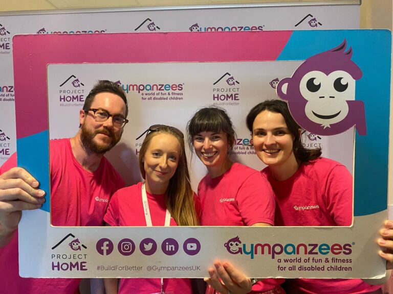 Four volunteers in pink t-shirts are smiling and looking through a Gympanzees frame