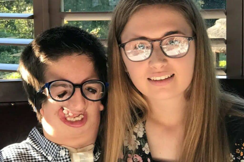Two siblings are smiling for a photo, one has short dark hair and glasses, the other has long blonde hair and glasses.