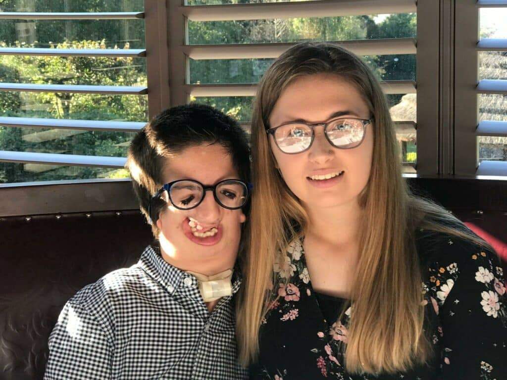 Two siblings are smiling for a photo, one has short dark hair and glasses, the other has long blonde hair and glasses.
