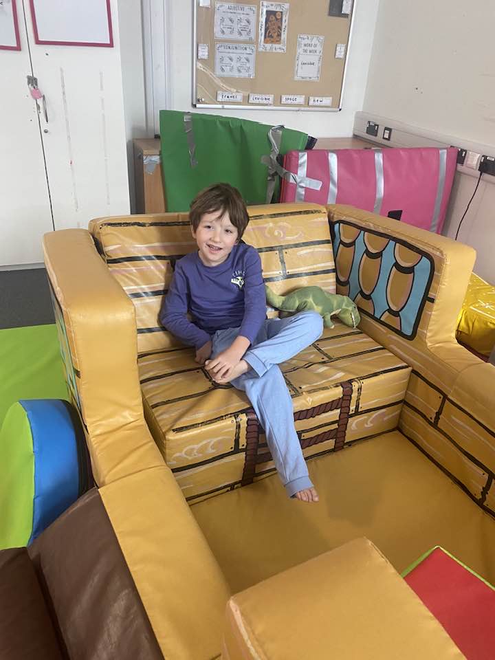 A child sits cross-legged in a soft play pirate ship, smiling and looking directly at the camera.